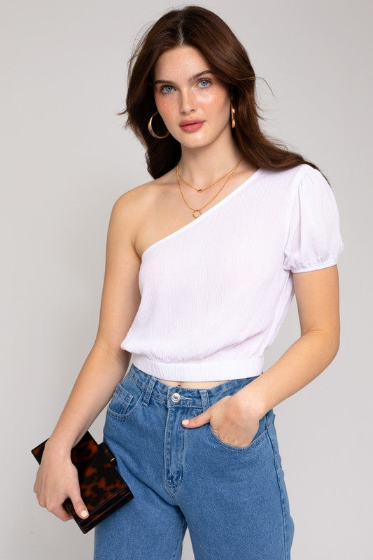 A person in a fashionable wardrobe featuring an asymmetrical design, wearing the S/S One Shoulder Top and jeans, holds a small rectangular purse while posing against a plain background.