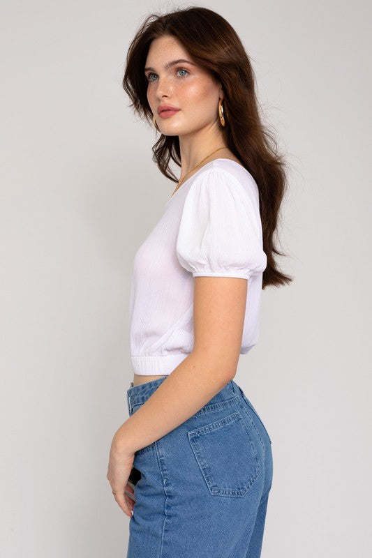 A person in a fashionable wardrobe featuring an asymmetrical design, wearing the S/S One Shoulder Top and jeans, holds a small rectangular purse while posing against a plain background.