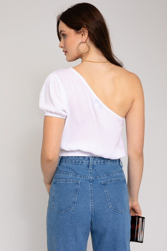 A person in a fashionable wardrobe featuring an asymmetrical design, wearing the S/S One Shoulder Top and jeans, holds a small rectangular purse while posing against a plain background.