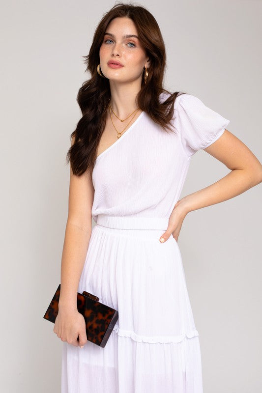 A person in a fashionable wardrobe featuring an asymmetrical design, wearing the S/S One Shoulder Top and jeans, holds a small rectangular purse while posing against a plain background.