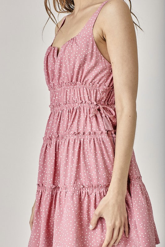 A person wearing the Side Tassel Strap Tiered Polka Dot Dress in pink, with a pattern of polka dots and adorned with side tassel straps and tiered ruffles, is gazing downward while slightly turning to the side.