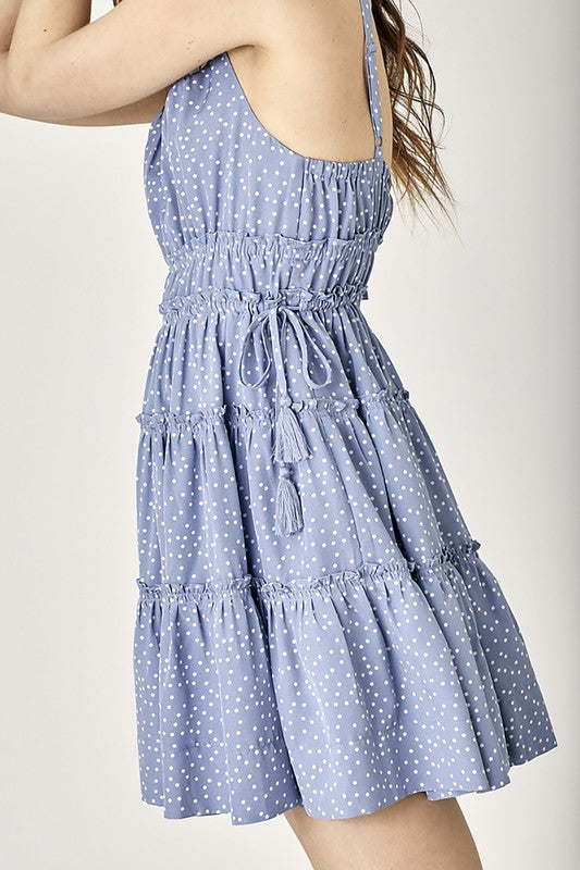 Against a simple background, a woman showcases boho flair in the sleeveless Side Tassel Strap Tiered Polka Dot Dress. This blue sundress is highlighted by its ruffled skirt and charming tassel-adorned straps, adding an extra whimsical touch.