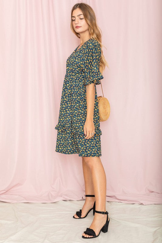 A woman wearing a Floral V Neck Ruffle Dress holds a wicker handbag, standing in a room decorated with a plant and partially visible furniture in the background.