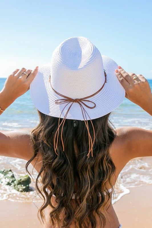 A woman holding onto her Beaded Belt Straw Panama Fedora Hat while standing on a beach.