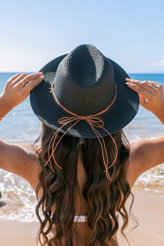 A woman holding onto her Beaded Belt Straw Panama Fedora Hat while standing on a beach.
