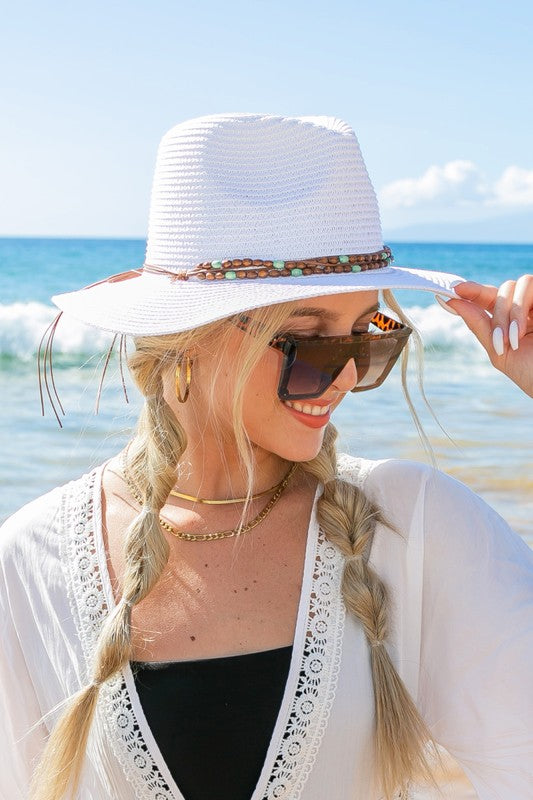 A woman holding onto her Beaded Belt Straw Panama Fedora Hat while standing on a beach.