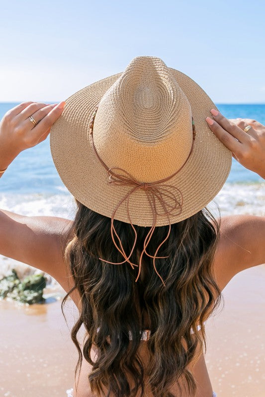 A woman holding onto her Beaded Belt Straw Panama Fedora Hat while standing on a beach.