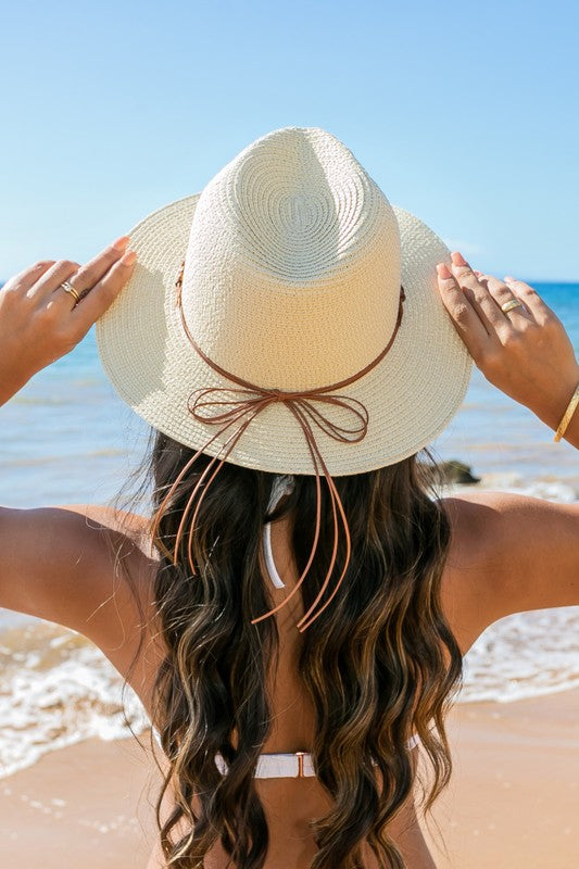 A woman holding onto her Beaded Belt Straw Panama Fedora Hat while standing on a beach.