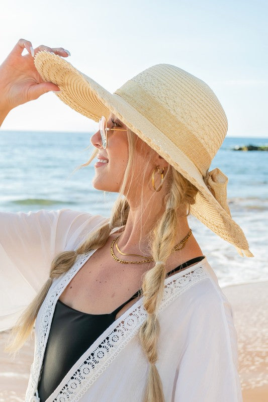 A woman holding her Scallop Edge Bow Accent Sunhat while standing on a beach.