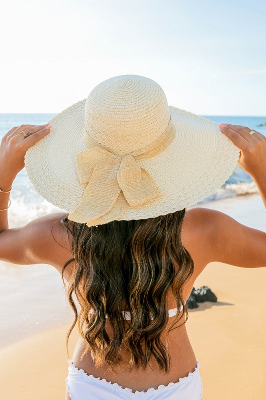 A woman holding her Scallop Edge Bow Accent Sunhat while standing on a beach.