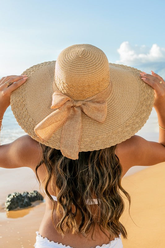 A woman holding her Scallop Edge Bow Accent Sunhat while standing on a beach.
