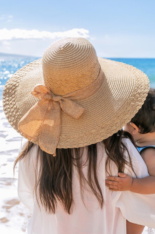 A woman holding her Scallop Edge Bow Accent Sunhat while standing on a beach.