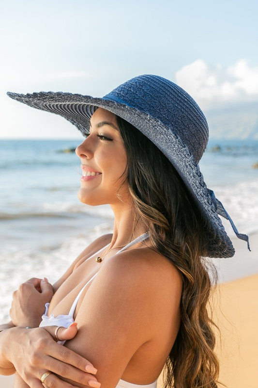 A woman holding her Scallop Edge Bow Accent Sunhat while standing on a beach.