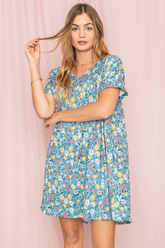 A woman stands against a pink backdrop, elegantly holding a strand of her hair while wearing the V-Neck Floral Baby Doll Dress.