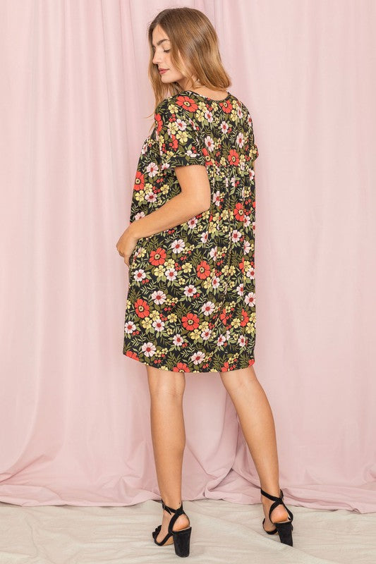 A woman stands against a pink backdrop, elegantly holding a strand of her hair while wearing the V-Neck Floral Baby Doll Dress.