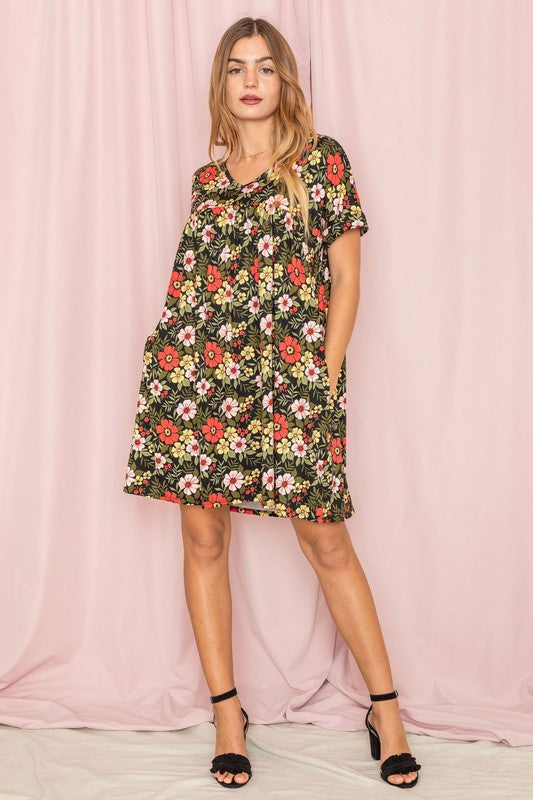 A woman stands against a pink backdrop, elegantly holding a strand of her hair while wearing the V-Neck Floral Baby Doll Dress.