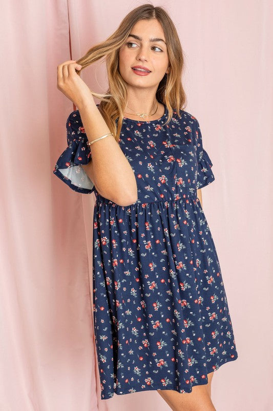 Wearing the Ditsy Floral Ruffle Chest Pocket Dress in navy, a woman poses gracefully in front of a pink curtain, gently touching her hair.