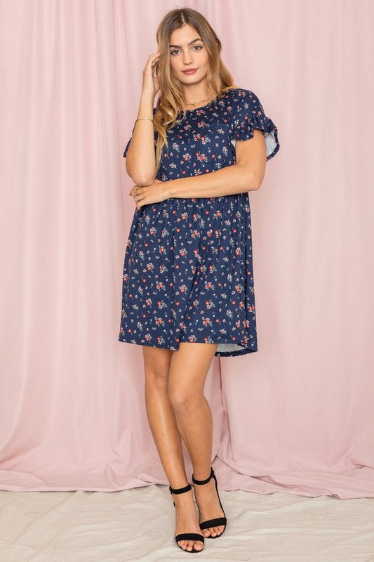 Wearing the Ditsy Floral Ruffle Chest Pocket Dress in navy, a woman poses gracefully in front of a pink curtain, gently touching her hair.