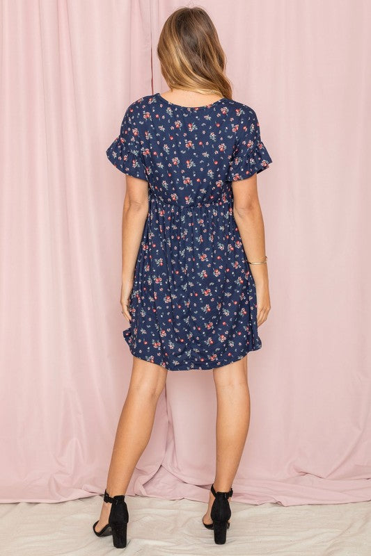 Wearing the Ditsy Floral Ruffle Chest Pocket Dress in navy, a woman poses gracefully in front of a pink curtain, gently touching her hair.