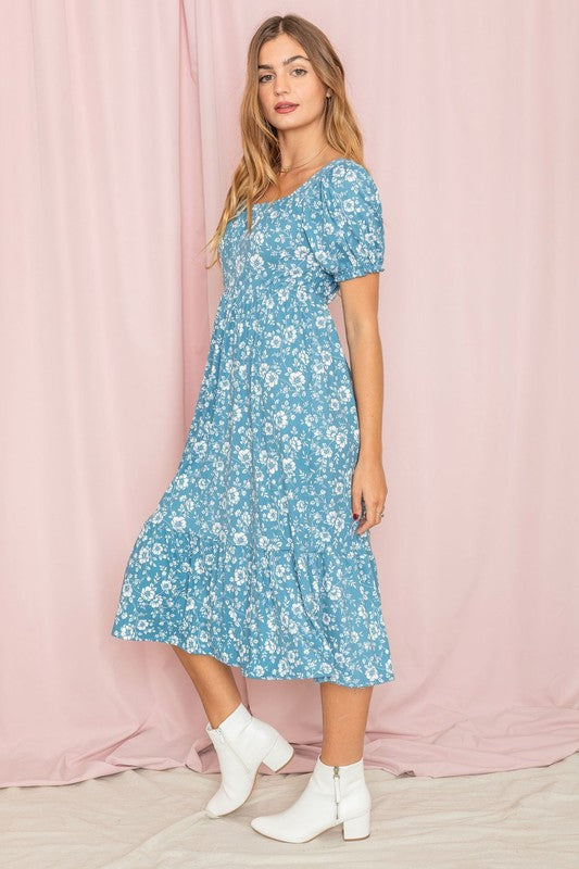 A woman wearing the Floral Square Neck Puff Sleeve Boho Dress is standing in front of a pink curtain, gently holding a strand of hair with one hand.