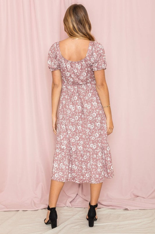 A woman wearing the Floral Square Neck Puff Sleeve Boho Dress is standing in front of a pink curtain, gently holding a strand of hair with one hand.