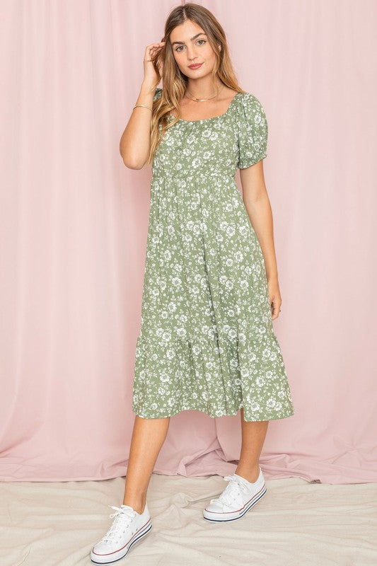 A woman wearing the Floral Square Neck Puff Sleeve Boho Dress is standing in front of a pink curtain, gently holding a strand of hair with one hand.