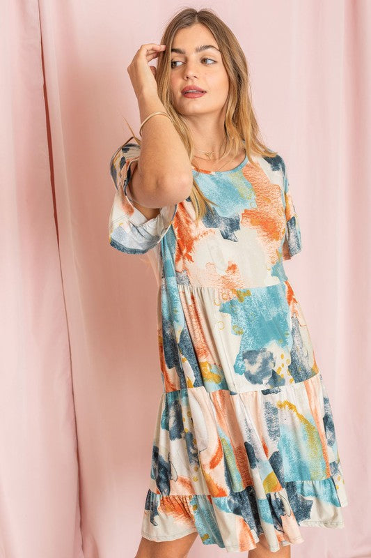 A woman in a vibrant Tie Dye Ruffle Triple Tiered Midi Dress with white ankle boots stands gracefully in front of a pale pink curtain.