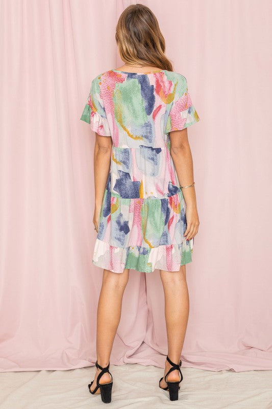 A woman in a vibrant Tie Dye Ruffle Triple Tiered Midi Dress with white ankle boots stands gracefully in front of a pale pink curtain.