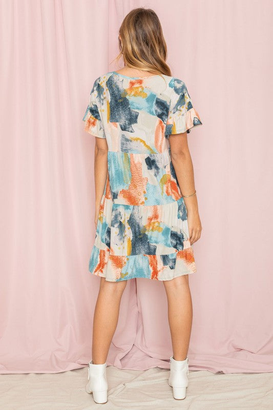 A person wearing a vibrant tie-dye ruffle midi dress and white ankle boots stands in front of a pink backdrop.