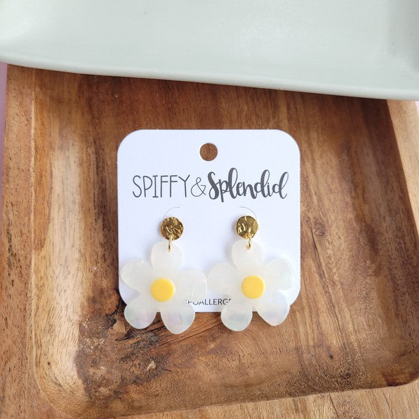 A pair of Daisy Dangles earrings, fashioned into charming flower shapes with yellow centers, rests elegantly on a white surface. These hypoallergenic stainless steel earrings are enhanced with 18K gold-plating and are set beside a rustic wooden board and a vibrant green plant.