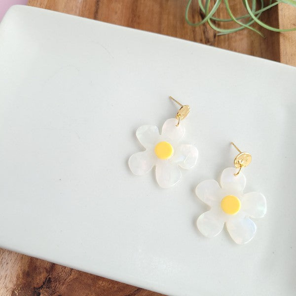 A pair of Daisy Dangles earrings, fashioned into charming flower shapes with yellow centers, rests elegantly on a white surface. These hypoallergenic stainless steel earrings are enhanced with 18K gold-plating and are set beside a rustic wooden board and a vibrant green plant.
