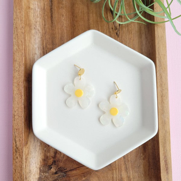 A pair of Daisy Dangles earrings, fashioned into charming flower shapes with yellow centers, rests elegantly on a white surface. These hypoallergenic stainless steel earrings are enhanced with 18K gold-plating and are set beside a rustic wooden board and a vibrant green plant.