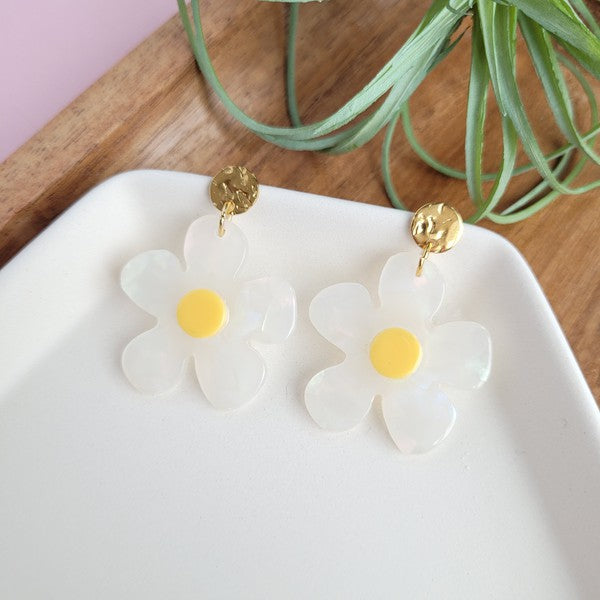 A pair of Daisy Dangles earrings, fashioned into charming flower shapes with yellow centers, rests elegantly on a white surface. These hypoallergenic stainless steel earrings are enhanced with 18K gold-plating and are set beside a rustic wooden board and a vibrant green plant.