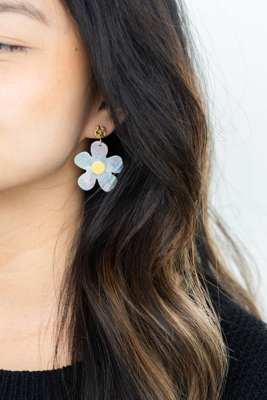 A pair of Daisy Dangles earrings, fashioned into charming flower shapes with yellow centers, rests elegantly on a white surface. These hypoallergenic stainless steel earrings are enhanced with 18K gold-plating and are set beside a rustic wooden board and a vibrant green plant.