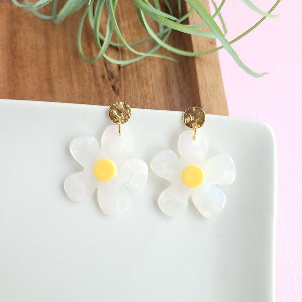 A pair of Daisy Dangles earrings, fashioned into charming flower shapes with yellow centers, rests elegantly on a white surface. These hypoallergenic stainless steel earrings are enhanced with 18K gold-plating and are set beside a rustic wooden board and a vibrant green plant.