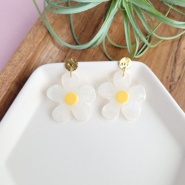 A pair of Daisy Dangles earrings, fashioned into charming flower shapes with yellow centers, rests elegantly on a white surface. These hypoallergenic stainless steel earrings are enhanced with 18K gold-plating and are set beside a rustic wooden board and a vibrant green plant.