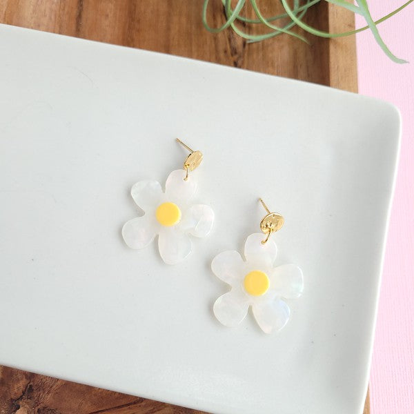 A pair of Daisy Dangles earrings, fashioned into charming flower shapes with yellow centers, rests elegantly on a white surface. These hypoallergenic stainless steel earrings are enhanced with 18K gold-plating and are set beside a rustic wooden board and a vibrant green plant.