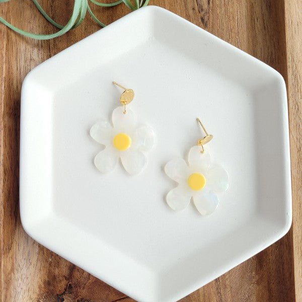 A pair of Daisy Dangles earrings, fashioned into charming flower shapes with yellow centers, rests elegantly on a white surface. These hypoallergenic stainless steel earrings are enhanced with 18K gold-plating and are set beside a rustic wooden board and a vibrant green plant.