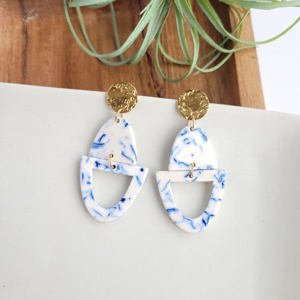 Athena Greek Goddess collection features stunning white and blue marbled earrings with gold accents, elegantly arranged on a hexagonal plate set on a wooden tray. These earrings are crafted with hypoallergenic stainless steel, perfect for sensitive ears.