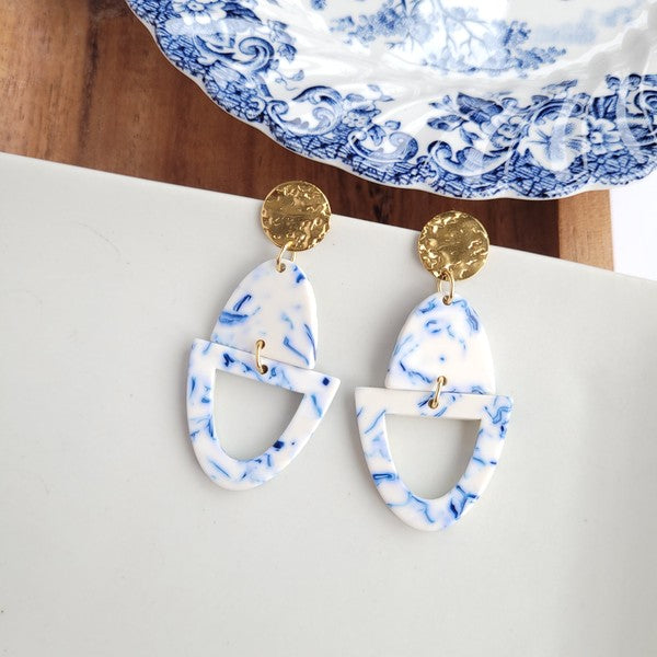 Athena Greek Goddess collection features stunning white and blue marbled earrings with gold accents, elegantly arranged on a hexagonal plate set on a wooden tray. These earrings are crafted with hypoallergenic stainless steel, perfect for sensitive ears.