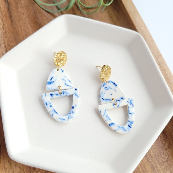 Athena Greek Goddess collection features stunning white and blue marbled earrings with gold accents, elegantly arranged on a hexagonal plate set on a wooden tray. These earrings are crafted with hypoallergenic stainless steel, perfect for sensitive ears.