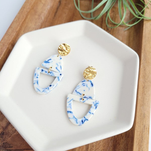 Athena Greek Goddess collection features stunning white and blue marbled earrings with gold accents, elegantly arranged on a hexagonal plate set on a wooden tray. These earrings are crafted with hypoallergenic stainless steel, perfect for sensitive ears.