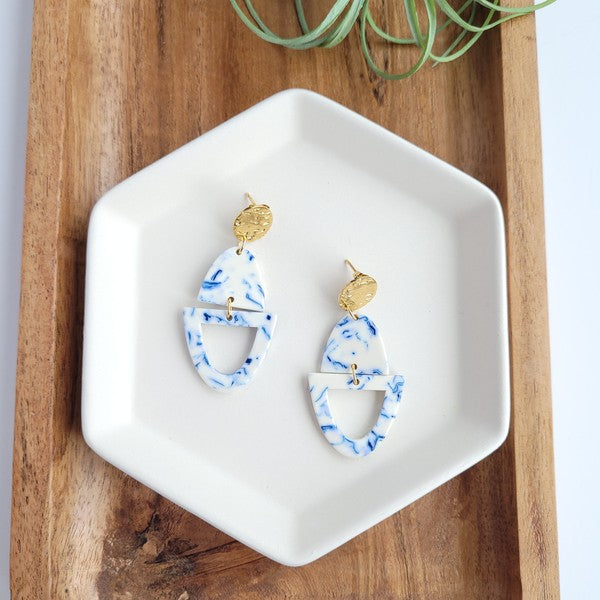 Athena Greek Goddess collection features stunning white and blue marbled earrings with gold accents, elegantly arranged on a hexagonal plate set on a wooden tray. These earrings are crafted with hypoallergenic stainless steel, perfect for sensitive ears.