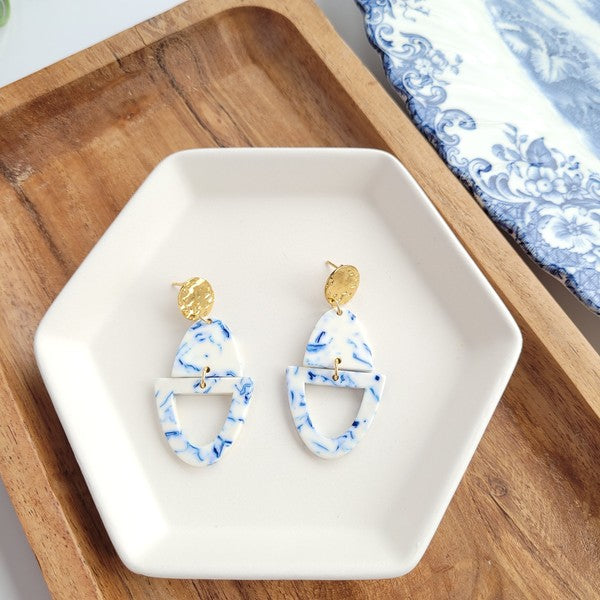 Athena Greek Goddess collection features stunning white and blue marbled earrings with gold accents, elegantly arranged on a hexagonal plate set on a wooden tray. These earrings are crafted with hypoallergenic stainless steel, perfect for sensitive ears.