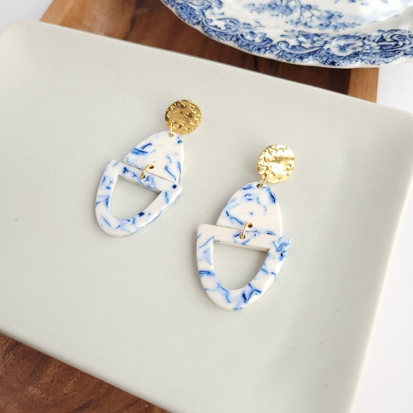 Athena Greek Goddess collection features stunning white and blue marbled earrings with gold accents, elegantly arranged on a hexagonal plate set on a wooden tray. These earrings are crafted with hypoallergenic stainless steel, perfect for sensitive ears.