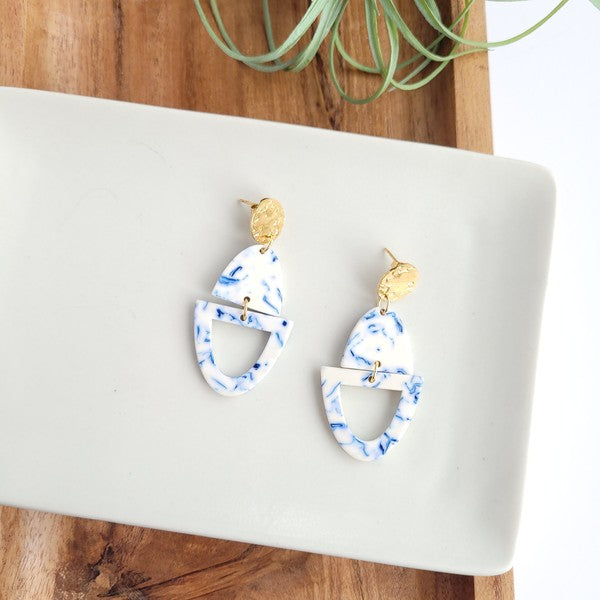 Athena Greek Goddess collection features stunning white and blue marbled earrings with gold accents, elegantly arranged on a hexagonal plate set on a wooden tray. These earrings are crafted with hypoallergenic stainless steel, perfect for sensitive ears.