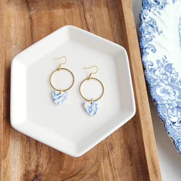 From the Greek Goddess collection, the Iris Earrings - Greek Goddess Blue feature gold hoops and heart-shaped blue and white pendants, elegantly displayed on a hexagonal white dish atop a wooden surface. Made from hypoallergenic stainless steel, they combine both style and comfort seamlessly.