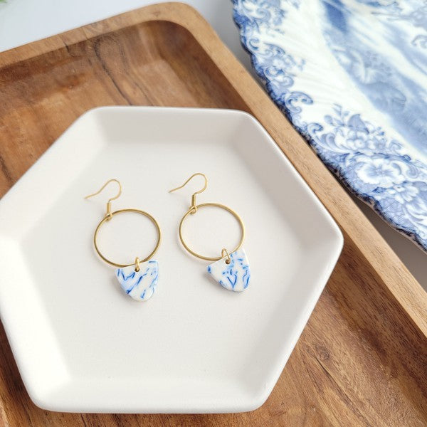 From the Greek Goddess collection, the Iris Earrings - Greek Goddess Blue feature gold hoops and heart-shaped blue and white pendants, elegantly displayed on a hexagonal white dish atop a wooden surface. Made from hypoallergenic stainless steel, they combine both style and comfort seamlessly.