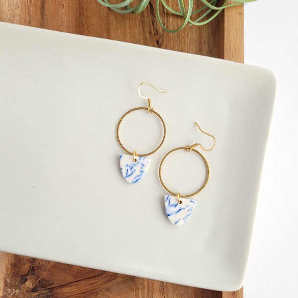 From the Greek Goddess collection, the Iris Earrings - Greek Goddess Blue feature gold hoops and heart-shaped blue and white pendants, elegantly displayed on a hexagonal white dish atop a wooden surface. Made from hypoallergenic stainless steel, they combine both style and comfort seamlessly.