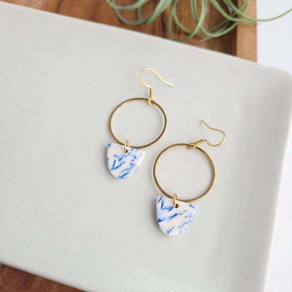 From the Greek Goddess collection, the Iris Earrings - Greek Goddess Blue feature gold hoops and heart-shaped blue and white pendants, elegantly displayed on a hexagonal white dish atop a wooden surface. Made from hypoallergenic stainless steel, they combine both style and comfort seamlessly.